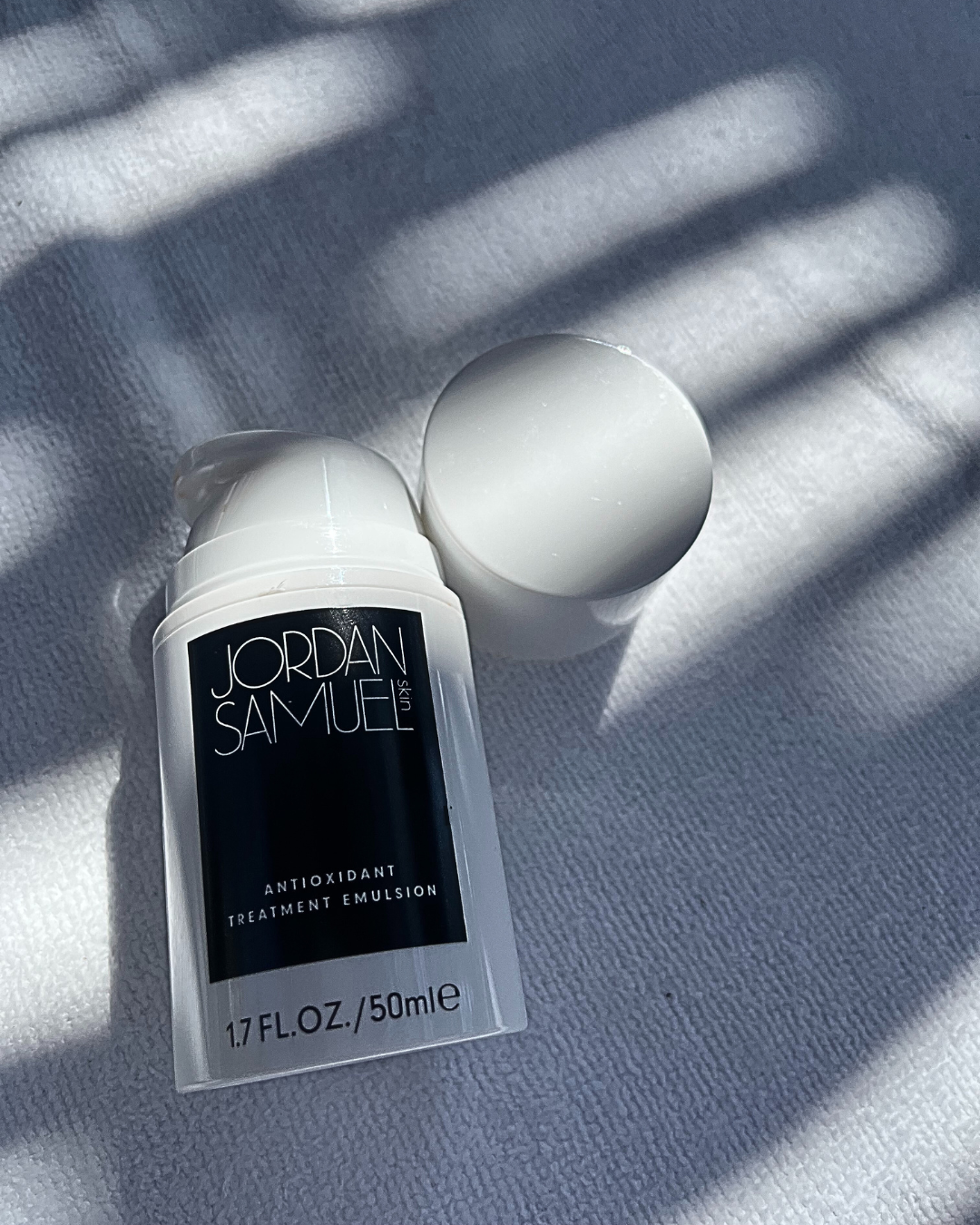 Antioxidant Treatment Emulsion lays on a white towel as sun streams through a pergola, casting it in shadows. 