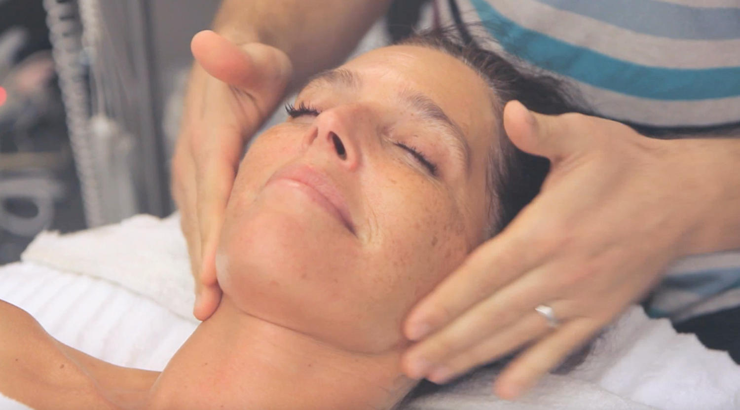 Woman lying on a facial bed. Esthetician's hands are seen performing facial massage at the woman's jawline. 