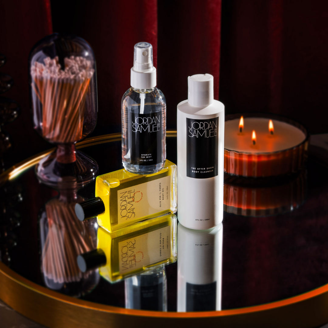 Olio Per Il Corpo, Hydrate the Mist, and The After Show Body Cleanser are clustered in the center of the image, resting on a gold-rimmed mirror tray. A red velvet curtain hangs in the background. 