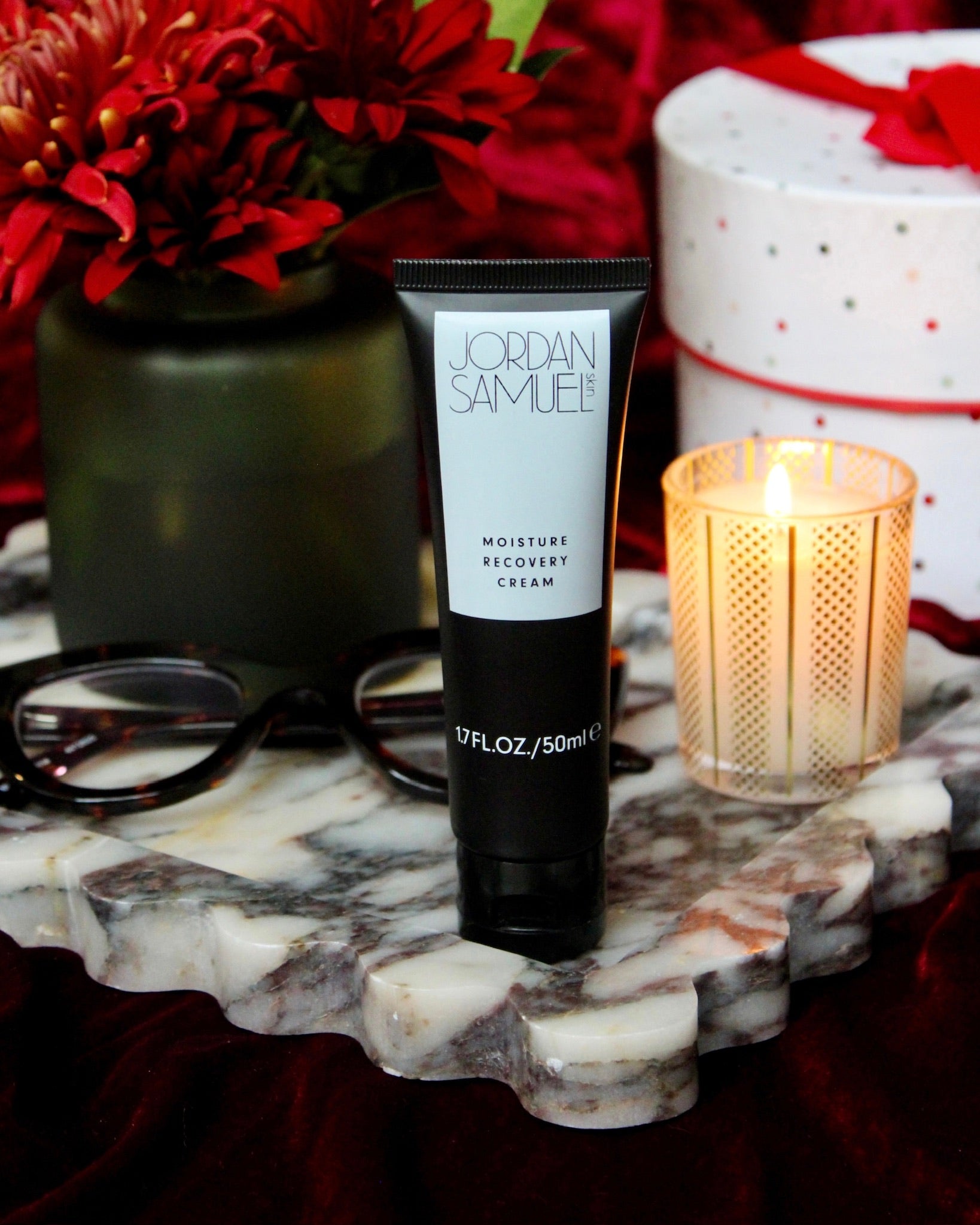 Moisture Recovery Cream arranged with eye glasses, lit candle, red and white gift box, and a red flower in dark green vase sit atop a white marble tray with scalloped edges.