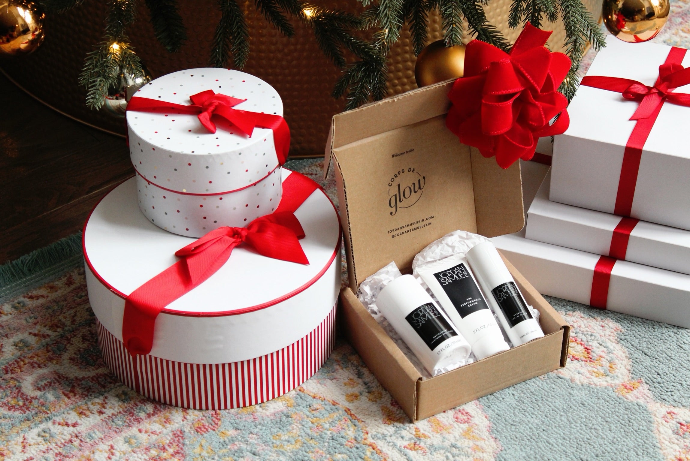 A Jordan Samuel shipping box sits below a Christmas tree amongst red and white gift boxes tied in red ribbon. The shipping box lid is ajar, showcasing Antioxidant Treatmetn Emulsion, The Performance Cream, adn Hydrate Facial Serum. 